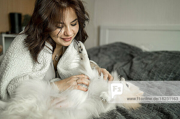 Happy young woman stroking Pomeranian dog on bed at home