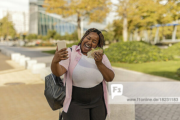 Fröhliche junge Frau mit Sandwich  die ein Selfie mit dem Smartphone macht