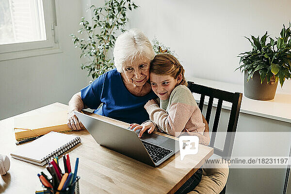 Lächelnde Großmutter hilft Enkelin beim Lernen per Laptop auf dem heimischen Tisch