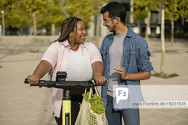 Happy couple with electric push scooter and bag of vegetables standing at footpath
