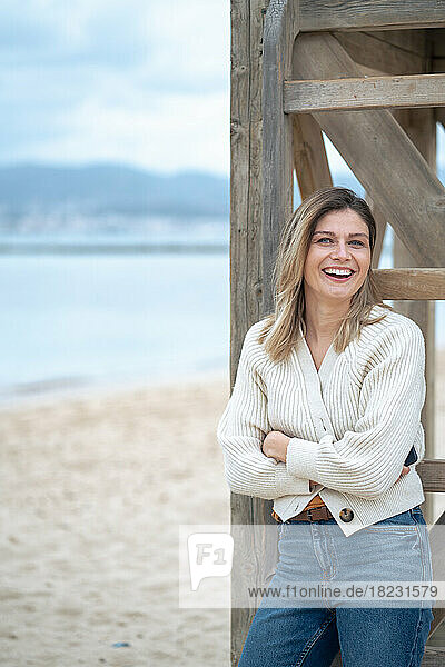 Happy young woman with arms crossed leaning on wooden lifeguard hut at beach