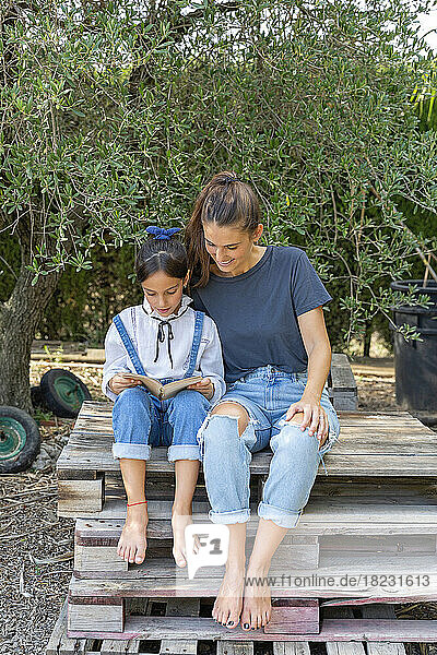 Girl reading book sitting by mother on wooden pallet