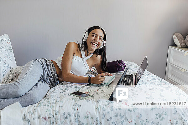 Happy girl wearing headphones and making notes with gadgets in bedroom