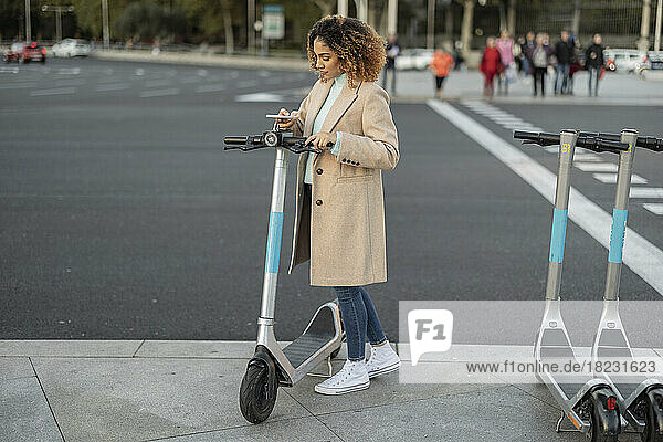 Young woman renting electric push scooter through smart phone on footpath