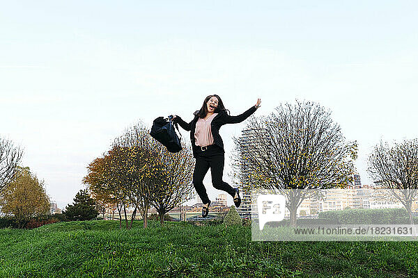 Happy young woman holding bag jumping in park