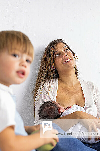 Contemplative mother breastfeeding newborn daughter by son at home