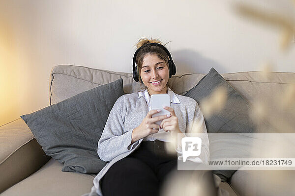 Happy woman wearing wireless headphones using smart phone on sofa at home