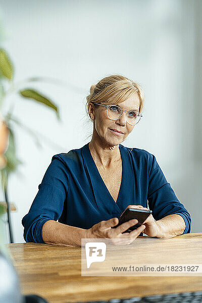 Thoughtful businesswoman with eyeglasses holding mobile phone at desk