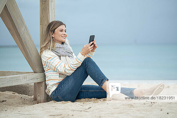 Smiling beautiful young woman using smart phone leaning on wood at beach