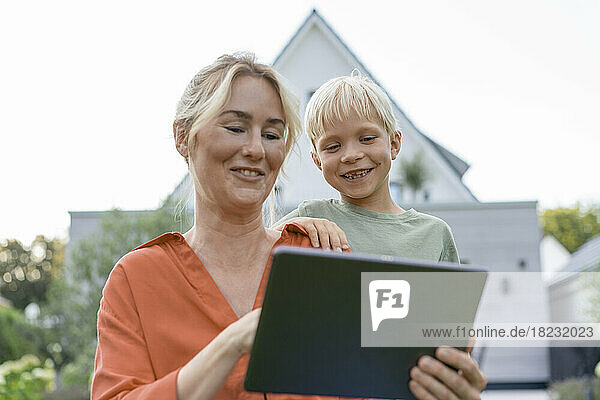 Smiling woman showing tablet PC to boy in front of house