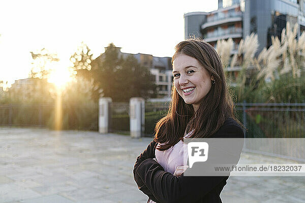 Happy young woman with arms crossed on footpath at sunset