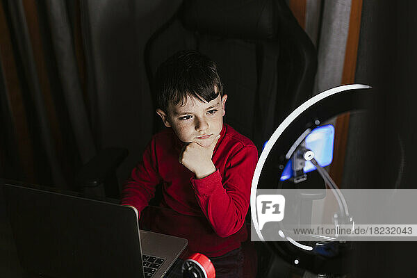 Nachdenklicher Junge sitzt mit Laptop und schaut im beleuchteten Ringlicht auf sein Mobiltelefon
