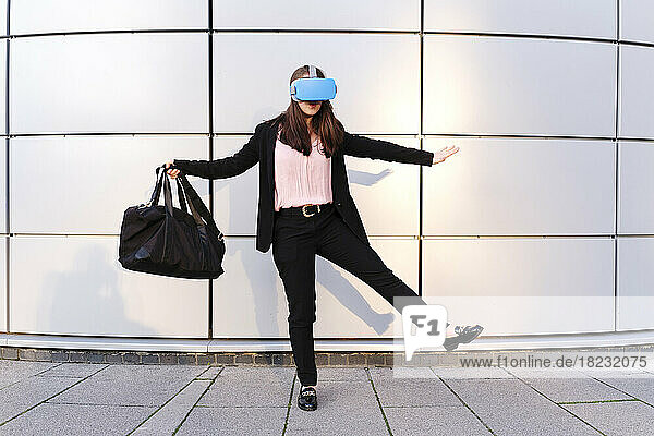 Young woman wearing virtual reality simulator holding bag in front of wall