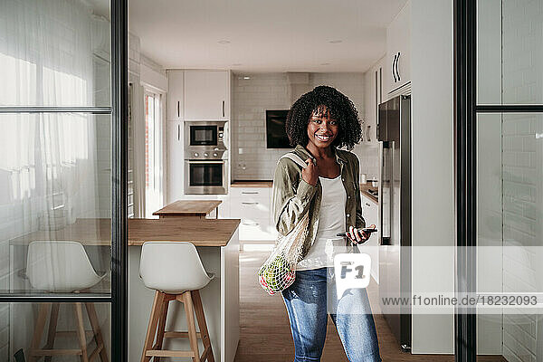 Smiling woman with mesh bag and smart phone standing at home