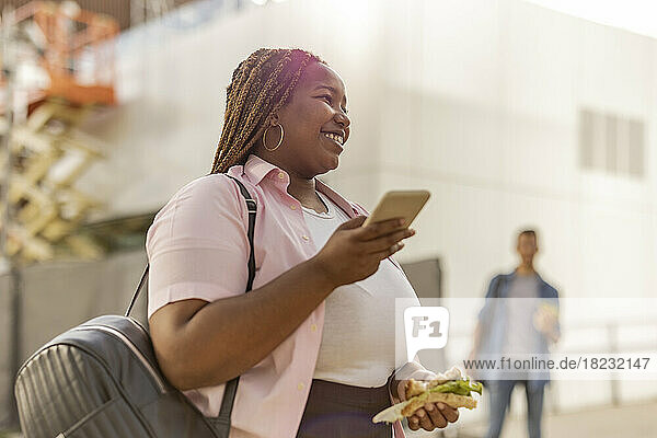 Smiling curvy woman with smart phone and sandwich