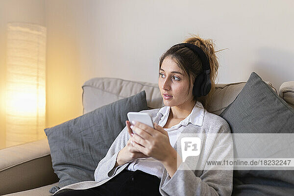 Nachdenkliche Frau mit Smartphone sitzt zu Hause auf dem Sofa