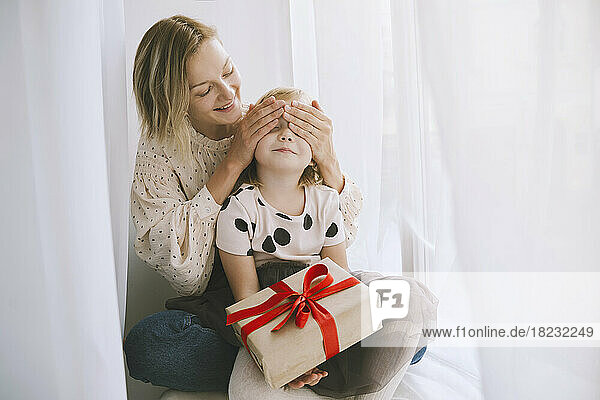 Mother covering eyes of daughter with Christmas present in front of curtain