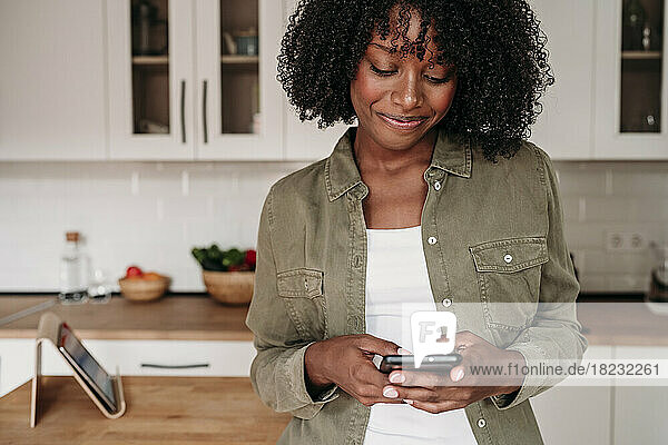 Lächelnde Frau  die zu Hause in der Küche Textnachrichten über ihr Smartphone sendet