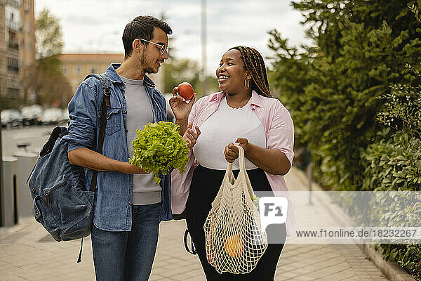 Happy woman showing vegetables to boyfriend standing at footpath