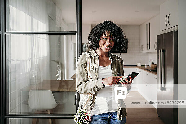 Junge Frau mit Smartphone und Netztasche steht zu Hause