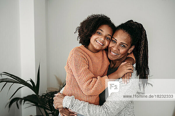 Happy woman embracing daughter in front of wall at home