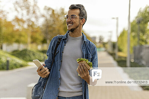 Cheerful young man with sandwich and smart phone standing on footpath