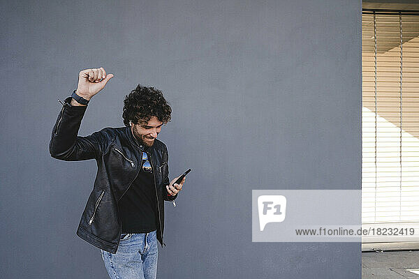 Glücklicher Mann mit erhobener Hand  der sein Mobiltelefon in der Hand hält und vor einer grauen Wand genießt
