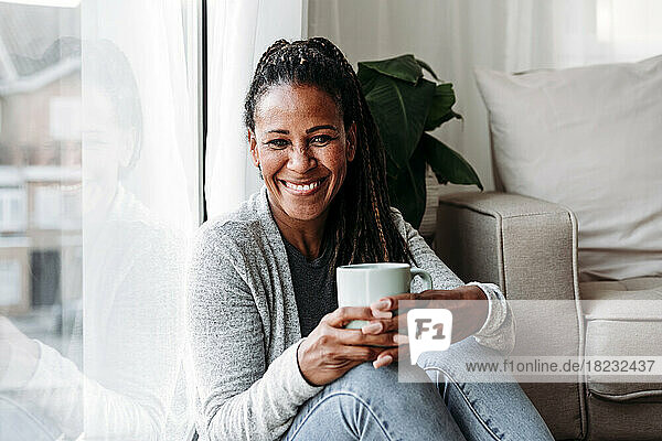 Glückliche reife Frau  die zu Hause mit einer Kaffeetasse am Fenster sitzt