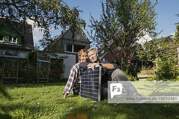 Smiling couple sitting with solar panel on grass in back yard