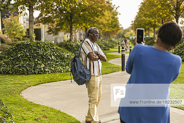 Frau fotografiert Mann  der mit verschränkten Armen im Park steht