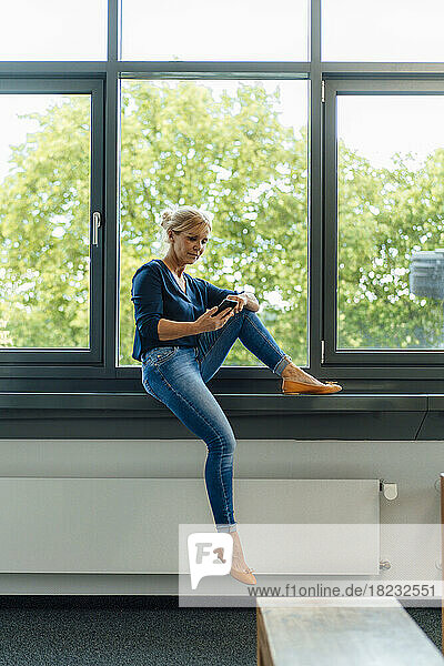 Businesswoman using smart phone sitting on window sill at office
