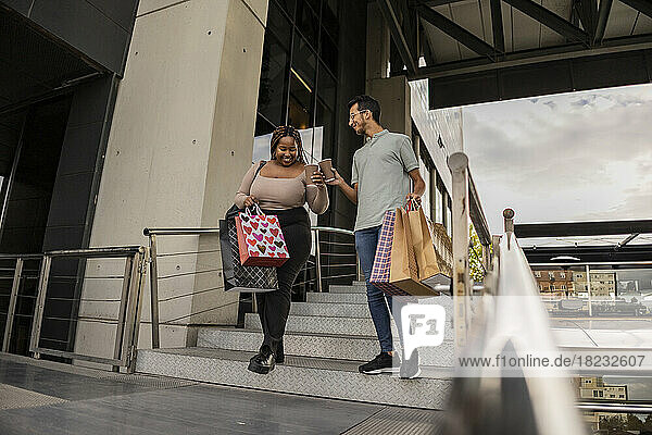 Happy young couple walking with shopping bags and toasting coffee cups on staircase