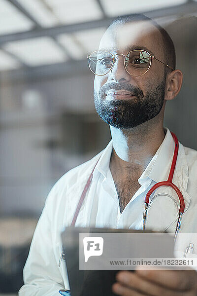 Smiling doctor holding tablet PC in hospital seen through glass