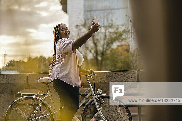 Smiling woman standing with bicycle and taking selfie through smart phone