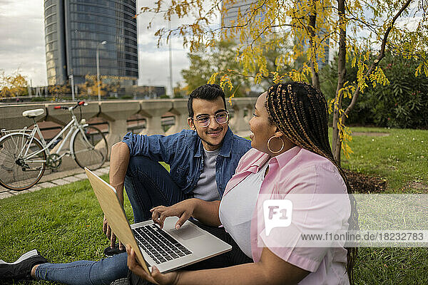 Smiling man and woman discussing and sitting with laptop on grass
