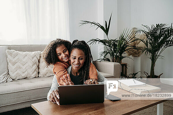 Glückliches Mädchen mit Mutter  die zu Hause im Wohnzimmer einen Laptop für Videoanrufe nutzt