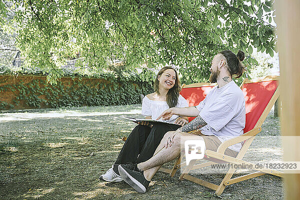 Happy couple with book sitting on deck chairs in backyard