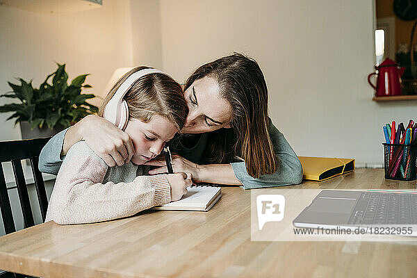 Mutter küsst Tochter  die zu Hause am Tisch Hausaufgaben macht