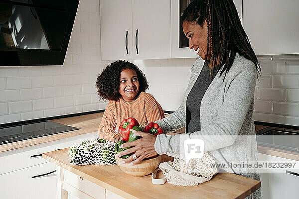 Lächelndes Mädchen mit Mutter  die zu Hause Gemüse auf dem Tisch hält