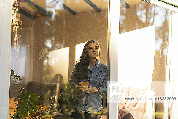Reife Frau mit Kaffeetasse und Blick aus dem Fenster eines Hauses