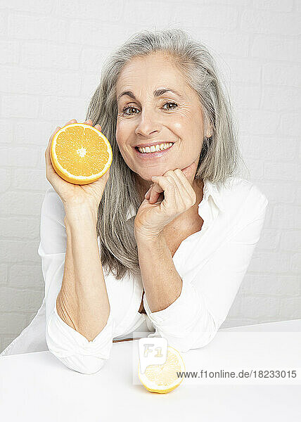 Happy mature woman with oranges leaning at table in front of wall