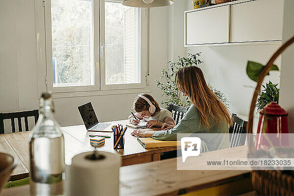 Mother looking at daughter doing homework sitting with laptop at table