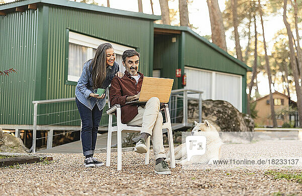 Lächelndes älteres Paar mit Hund und Laptop vor grünem Gebäude