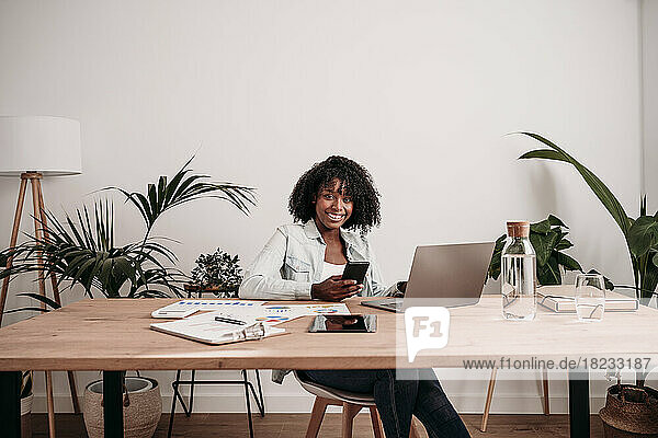 Young freelancer with smart phone and laptop on desk sitting at home office