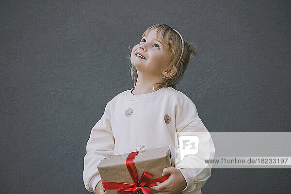 Lächelndes Mädchen schaut nach oben und hält eine Geschenkbox vor der Wand