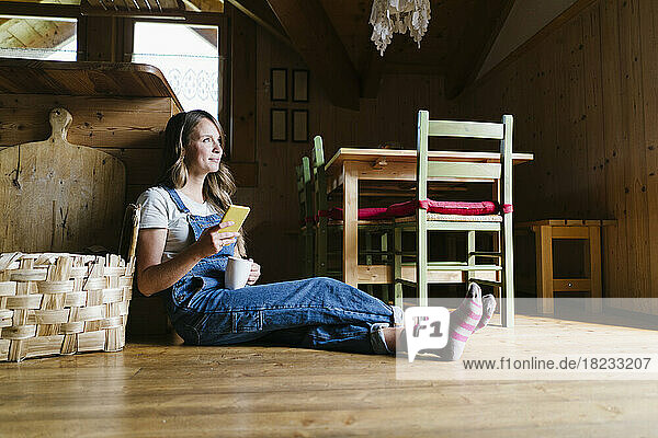 Thoughtful woman with coffee cup and smart phone sitting on floor