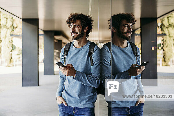 Smiling man with hand in pocket holding smart phone leaning on wall in corridor