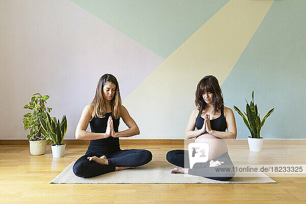 Pregnant woman meditating with yoga instructor sitting in front of wall at studio