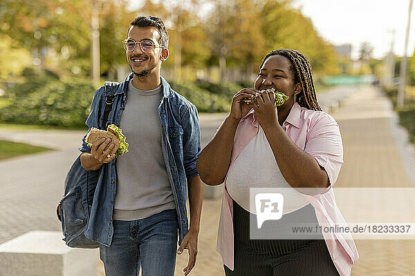 Glückliches junges Paar isst Sandwich und geht auf dem Fußweg
