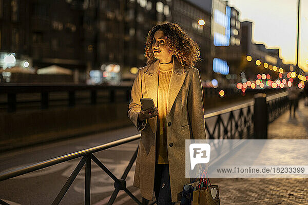 Frau hält Einkaufstüten und Mobiltelefon in der Abenddämmerung auf dem Fußweg
